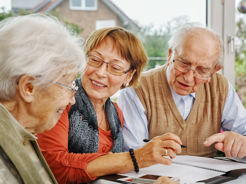 Three elderly people sitting together at a table reviewing a document inside a home.